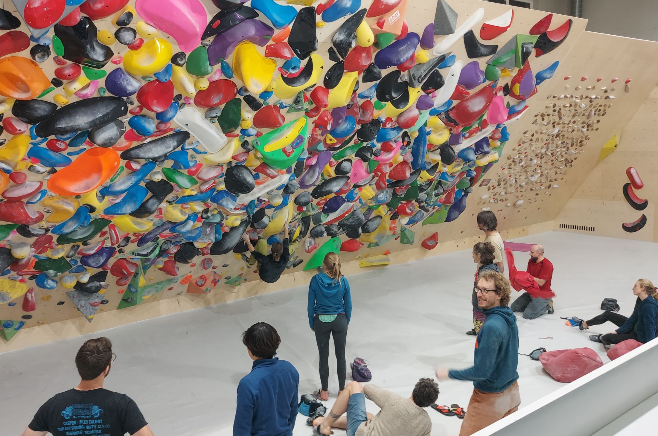 Bunte Spraywall mit vielen Griffen, vor der sich viele Boulderer unterhalten